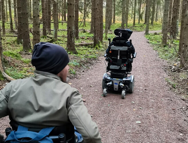 Kaksi henkilöä kulkee pyörätuoleilla Kumijoen ulkoilureitillä metsässä.