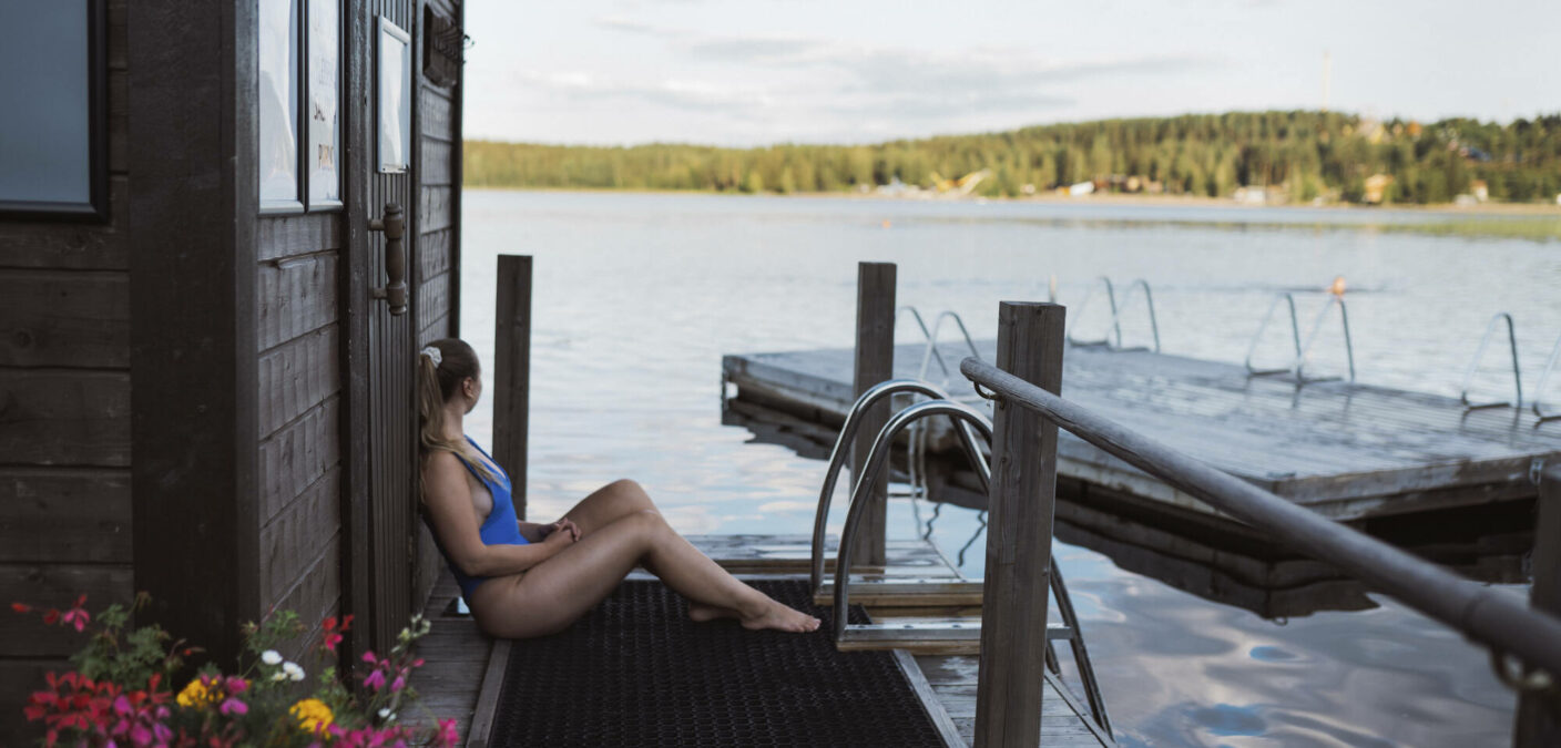 Nainen istuu rantasaunan laiturilla ja katselee järvelle päin.